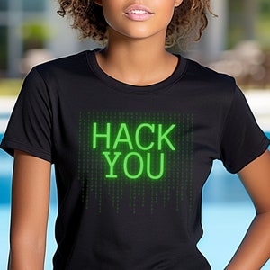 Op de afbeelding: Zwart T-shirt met een groene "HACK YOU" print op de voorkant. De print is gemaakt van een matrix van groene cijfers en letters.