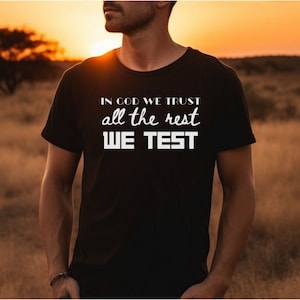 Può includere: Una maglietta nera con la scritta "In God We Trust all the rest We Test" stampata in bianco.