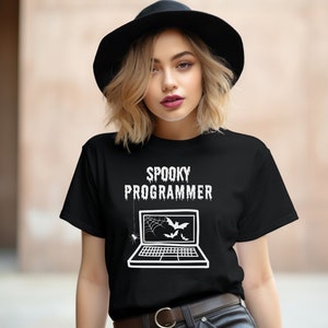 Könnte beinhalten: Schwarzes T-Shirt mit dem Text "SPOOKY PROGRAMMER" über einer Laptop-Grafik mit Spinnennetz und Fledermäusen. Die Person trägt einen schwarzen Hut.