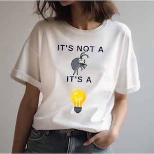 May include: White t-shirt with a graphic of a bug and a lightbulb. The text on the shirt reads "It's not a" above the bug and "It's a" below the lightbulb.