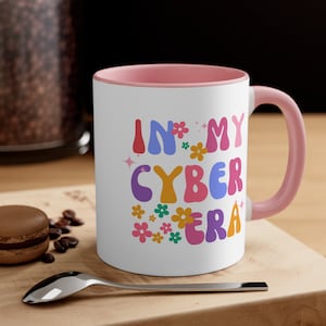 May include: White ceramic mug with a pink rim and handle. The mug has a colorful graphic that says "In My Cyber Era" with flowers and sparkles.
