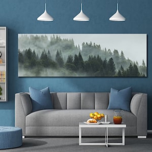 May include: A grey sofa with blue cushions sits in front of a large canvas print depicting a misty forest scene. A white coffee table with a bowl of fruit and a glass jug sits in front of the sofa.