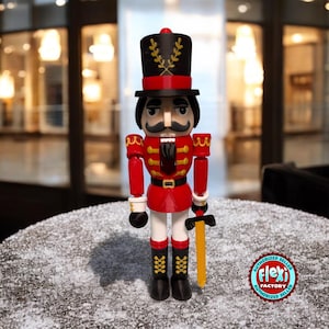 Puede incluir: Un cascanueces de juguete rojo y negro con una espada dorada. El cascanueces lleva un sombrero negro con ribetes dorados y un uniforme rojo y dorado. El cascanueces está de pie sobre una superficie blanca.