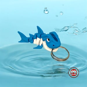 Puede incluir: Un llavero de tiburón azul y blanco con un llavero plateado. El tiburón está sujetando el llavero en su boca. El tiburón está flotando en una piscina de agua azul.