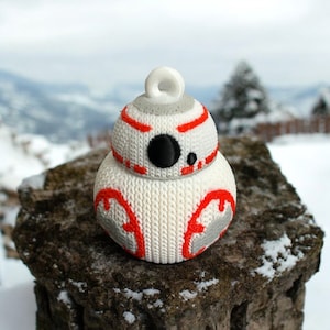 May include: A white and orange knitted ornament shaped like a droid with a single black eye. The ornament has a loop on top for hanging.