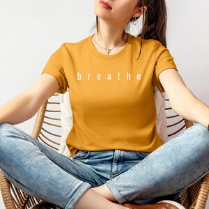 May include: A woman wearing a mustard yellow t-shirt with the word "breathe" printed in white on the front. She is sitting in a wicker chair wearing blue jeans.
