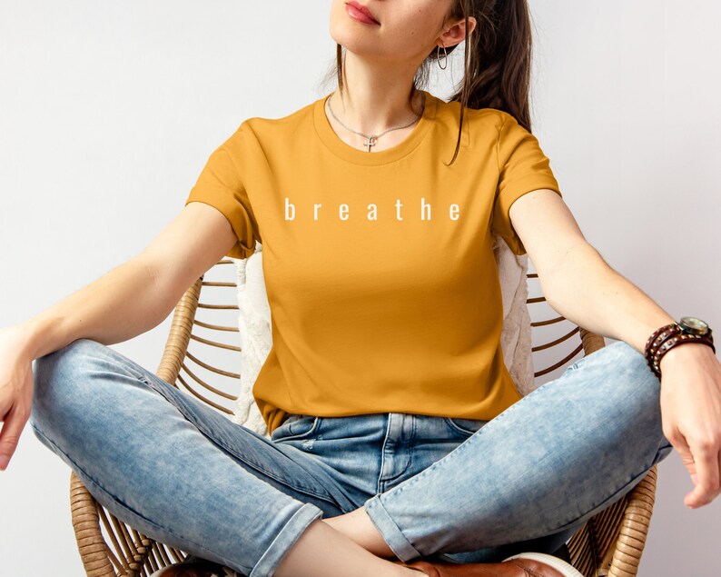 May include: A woman wearing a mustard yellow t-shirt with the word "breathe" printed in white on the front. She is sitting in a wicker chair wearing blue jeans.