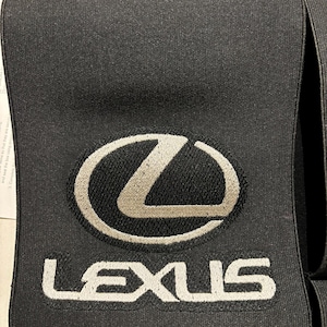 Puede incluir: Un accesorio de tela negra que presenta el logotipo plateado de Lexus bordado en la parte superior y la palabra LEXUS bordada en letras mayúsculas blancas debajo. El material tiene una apariencia texturizada y elástica con costuras visibles.