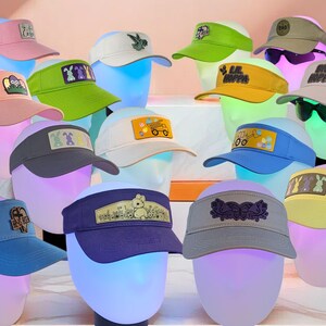 May include: A collection of 14 colorful visors with various embroidered designs, including bunnies, Easter eggs, and a truck. The visors are displayed on mannequins with a pink background.