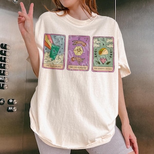 May include: A white t-shirt featuring three colourful illustrations of cards. The cards have the text "The Baja Blast", "The Crunchwrap", and "The Gorista Crunch".