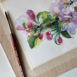 Hand holding a paintbrush near a horizontal watercolor painting of pink apple tree blossoms on white paper.