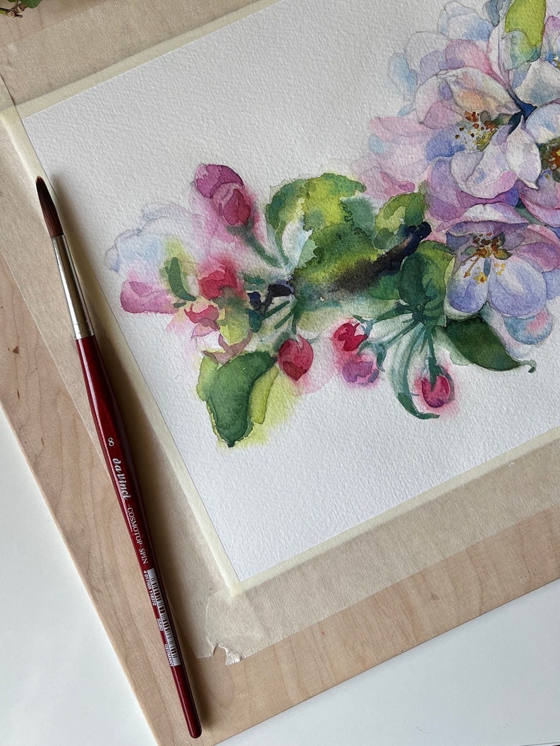 Hand holding a paintbrush near a horizontal watercolor painting of pink apple tree blossoms on white paper.