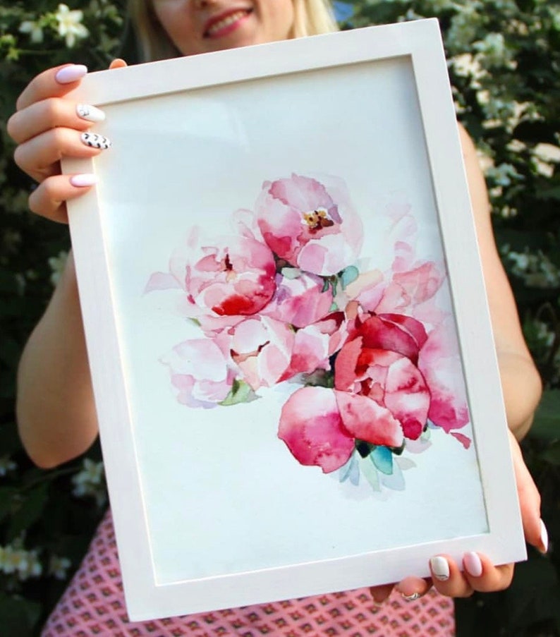 the girl holds a picture in a white frame in her hands. the painting depicts delicate pink roses in watercolor technique