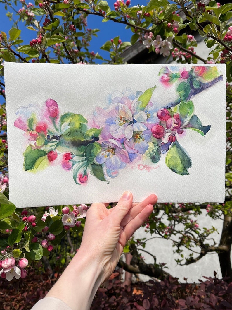 Original watercolor painting of blooming apple branches held against a real flowering tree and bright blue sky.