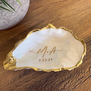 May include: A decorative oyster shell trinket dish with a gold-painted edge. The white interior features the initials "M+A" and the date "9.27.25" in gold script. The dish sits on a wooden surface.