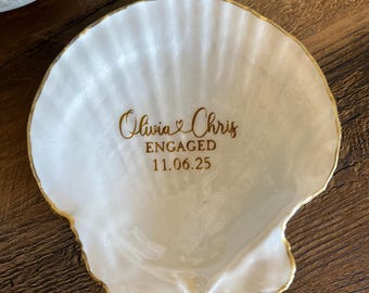 May include: White seashell-shaped trinket dish with gold trim. The dish has the words "Olivia & Chris ENGAGED 11.06.25" in gold script. The dish is on a wooden surface.
