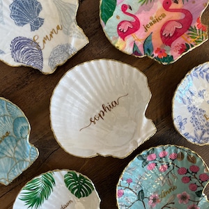 Puede incluir: Una colección de cuencos en forma de concha con borde dorado. Cada plato presenta un diseño único, incluyendo patrones florales, conchas marinas y nombres personalizados en escritura dorada. La paleta de colores incluye azul, rosa y verde.