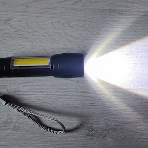 Op de afbeelding: Een zwarte zaklamp met een gele LED-lamp, aan en helder schijnend. De zaklamp heeft een polsbandje.