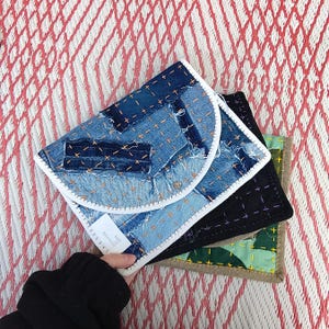 Pode incluir: Uma pilha de três bolsas de ganga feitas à mão. A bolsa superior é de ganga azul clara com costuras visíveis e uma borda branca. A bolsa do meio é preta e a inferior é verde com um padrão de camuflagem.