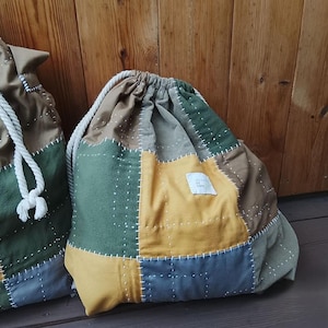 Puede incluir: Dos bolsas con cordón con un diseño de patchwork. Las bolsas están hechas de varias telas en tonos de verde, amarillo, marrón y azul. Las costuras blancas acentúan las costuras. Un cordón blanco de cuerda es visible en cada bolsa.