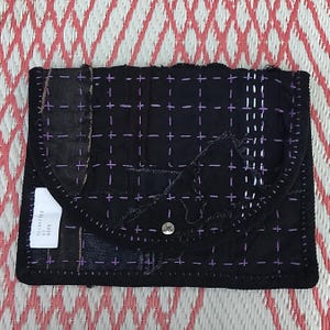 Op de afbeelding: Zwarte clutch van patchwork denim met paarse kruissteekdetails en een drukknoopsluiting. Een klein wit label leest "RECREATED BY HOPE".