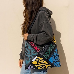 Könnte beinhalten: Eine große Tragetasche mit einem Patchwork-Design aus verschiedenfarbigen Stoffbahnen in Gelb, Blau, Grün, Rot und Violett, die jeweils mit komplexen, geschwungenen und gepunkteten Mustern verziert sind. Die Tasche hat einen schwarzen Trageriemen.