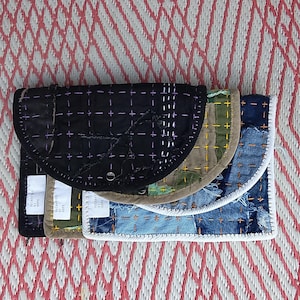 Op de afbeelding: Een stapel van drie handgemaakte stoffen portemonnees. De bovenste portemonnee is zwart met paarse stiksels, de middelste is camouflage en de onderste is van denim. Elke portemonnee heeft een gebogen flap en een label.