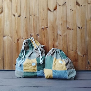Può includere: Due sacche con coulisse con un design patchwork. Le sacche presentano quadrati di tessuto verde, giallo, marrone e blu. Ogni sacca ha un cordoncino bianco e una piccola etichetta bianca. Le sacche sono su una superficie di legno.