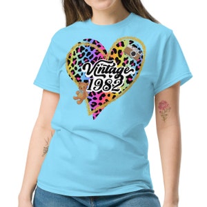 Peut inclure: Un t-shirt bleu clair avec un motif de cœur en imprimé léopard noir et arc-en-ciel. Le cœur a un contour de paillettes dorées et le texte "Vintage 1982" à l'intérieur. Un petit ours en peluche et un appareil photo vintage sont également inclus dans le design.