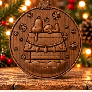 May include: A circular wooden Christmas ornament featuring a carved design of a dog sleeping on top of a doghouse. The scene includes engraved snowflakes and a string of lights draped across the roof. The text Digital CNC File - No Physical Product is visible at the bottom.