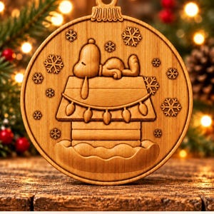 May include: A round wooden Christmas ornament featuring a carved Snoopy lying on top of a doghouse decorated with string lights, surrounded by snowflakes. The text at the bottom reads: Digital CNC File - No Physical Product.