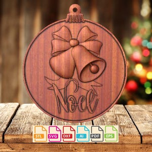 May include: A wooden Christmas bauble featuring a carved design of two bells, a bow, and the word "Noel". The ornament is brown and has a loop at the top for hanging. The background is a blurred image of a Christmas tree.