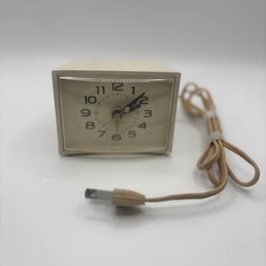 Puede incluir: Un reloj despertador eléctrico vintage de color crema con una esfera cuadrada y un cable de alimentación beige. La esfera del reloj tiene números y manecillas negras. Diseño clásico.