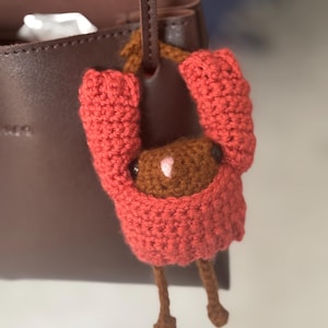 May include: A handmade coral-coloured crochet doll suspended from a brown leather handbag. The doll features a brown head and legs, with a small pink detail on its face. The doll's arms are raised, as if holding onto the handbag strap.