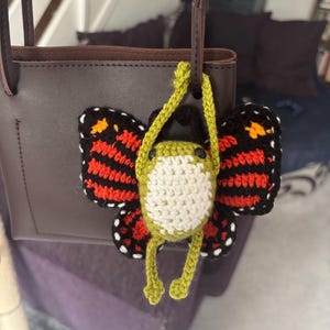 Könnte beinhalten: Eine braune Ledertasche mit einem gehäkelten Schmetterlingsanhänger. Der Schmetterling hat weiße, grüne, schwarze, orange und rote Details. Der Anhänger hängt am Riemen der Tasche.