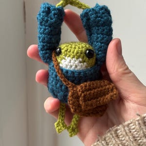 May include: A handmade crochet frog toy in teal blue, green, and brown. The frog has a small brown satchel and is holding onto a green vine. The frog's body is green and white, with black eyes.