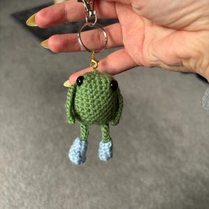 Puede incluir: Un llavero verde hecho a mano con ganchillo que presenta una pequeña criatura con ojos negros y botines azules. El llavero tiene un cierre plateado y un anillo dorado para sujetar las llaves. El cuerpo de la criatura es redondo con orejas pequeñas.