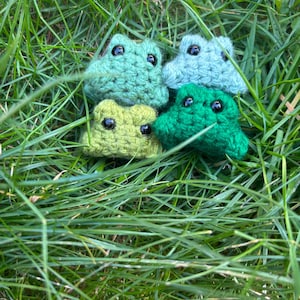 May include: Four crocheted frog figures in shades of green and blue, with black bead eyes, rest in a bed of green grass. The frogs vary in colour from light green to dark green and blue. The handmade amphibians are a whimsical craft.