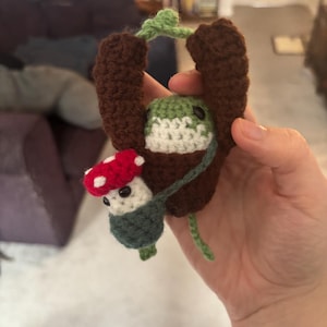 May include: A handmade crochet mushroom and log-shaped toy. The mushroom has a red cap with white spots, a white stem, and a green base. The log is brown with green leaves. The toy is held in a hand.
