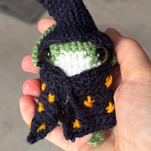 Può includere: Una rana all'uncinetto fatta a mano, che indossa un cappello e una veste da mago. La rana è verde e bianca con occhi neri a bottone. La veste blu scuro è decorata con forme di stella gialla. Un oggetto artigianale stravagante.