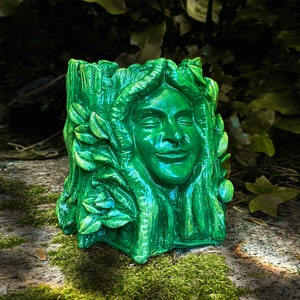 May include: A green ceramic planter shaped like a woman's face with leaves and vines. The planter has a smiling expression and is made to look like a tree trunk.
