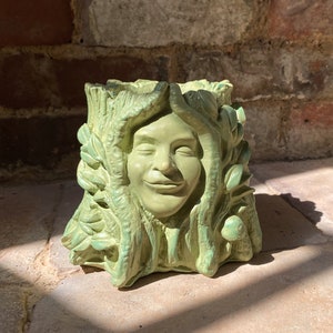 May include: A green ceramic planter shaped like a face with leaves and vines. The face has a smiling expression.