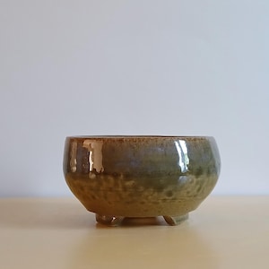May include: A small, handcrafted ceramic bowl with a rounded shape and three small feet. The bowl has a mottled glaze in shades of brown and green, giving it a rustic appearance. The interior is smooth and reflective.