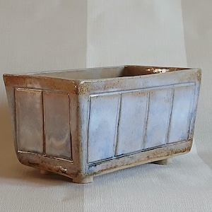 May include: Rectangular ceramic planter with a light blue and brown glaze. The planter has vertical rectangular panels on the sides and sits on small feet. The edges have a brown trim.