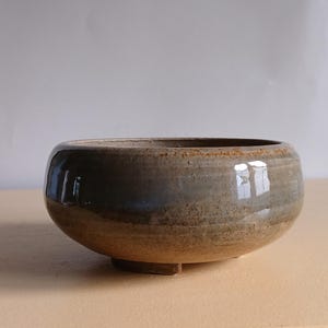 May include: A handmade ceramic bowl with a wide, rounded form. The bowl has a speckled, earthy colour scheme with shades of brown, grey, and tan. The exterior has a glossy finish, and the base has a small, rectangular foot.