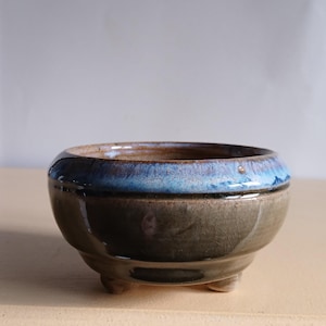 May include: A small, ceramic bonsai pot with a rounded shape and three small feet. The pot has a dark, earthy colour with a blue and brown glazed rim. The interior is a lighter brown.