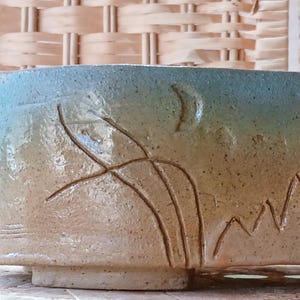 May include: A handmade ceramic bowl with a light brown base and a gradient of blue to tan on the upper portion. The bowl features etched designs of a crescent moon, plants, and mountains.
