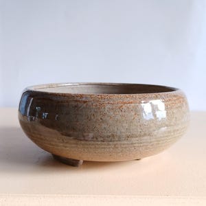 May include: A round, low ceramic planter with a speckled, earthy brown glaze. The planter has a wide opening and sits on three small feet. The interior is a lighter tan colour, and the rim has a darker, textured band.