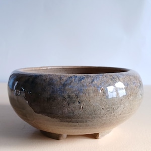 May include: A handmade, round ceramic planter with a wide opening and a low profile. The planter is a mottled brown with blue and grey accents and sits on three small feet. The glazed surface reflects light, adding a subtle sheen.