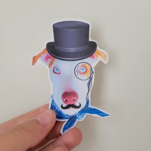 Puede incluir: Un perro blanco con un pañuelo azul y un sombrero de copa negro con un monóculo. El perro tiene la nariz rosa y los ojos azules.